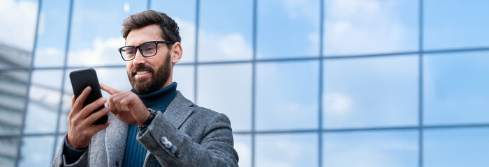 Successful businessman using mobile cellphone phone app, business chat for trade work outdoors. Mature man holding smartphone standing in city skyscrapers building on background. Banner, copy space