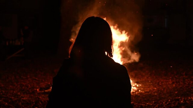 Back lit silhouette of adult female person looking on bonfire pyre at night, pyromania concept, 4K slow motion