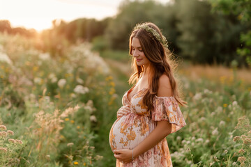 Pregnant woman standing in a field with sunlight and flowers, smiling in a summer dress outdoors
