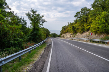 Road landscape forest, mountains and sea on the horizon. Empty long mountain road to the horizon on a sunny summer day with dramatic cloudy sky. Highway turn panoramic view.