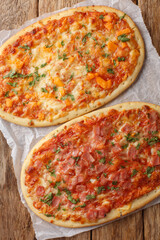 Italian oval pizza focaccia with cheese, ham and tomato sauce close-up on parchment on wooden table. Vertical top view from above