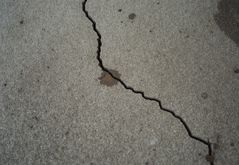 Close-up view of a jagged crack running diagonally across a textured concrete surface, revealing structural damage and weathering effects.
