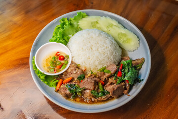 Stir-fried beef with basil in a blue plate with fish sauce, chili, cucumber and lettuce on a wooden table.