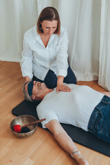 A man lying on a mat with a blindfold, practicing breathing exercises while a woman acts as a...