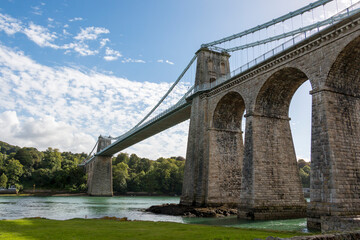 Fototapeta premium Menai suspension bridge. A stone arch bridge spans a river under a bright, partly cloudy sky.