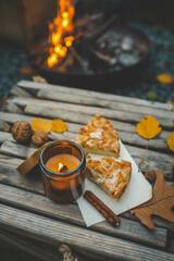 Close-up of a lit candle in an amber glass jar, two slices of apple pie, and a cinnamon stick on a rustic wooden table. Autumn leaves and walnuts enhance the cozy atmosphere near a warm campfire