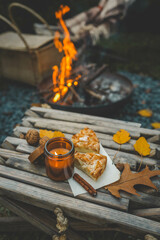 Close-up of a lit candle in an amber glass jar, two slices of apple pie, and a cinnamon stick on a rustic wooden table. Autumn leaves and walnuts enhance the cozy atmosphere near a warm campfire