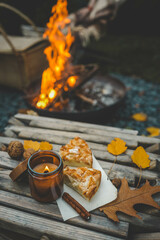 Close-up of a lit candle in an amber glass jar, two slices of apple pie, and a cinnamon stick on a rustic wooden table. Autumn leaves and walnuts enhance the cozy atmosphere near a warm campfire