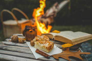 Rustic autumn scene with a piece of apple pie, walnuts, and cinnamon sticks on a wooden table near a warm campfire