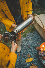 Close-up people pouring hot drinks from thermos into metal mugs next to campfire. Cozy autumn scene with warm clothing, a woven basket, and the glow of the fire, creating a warm 