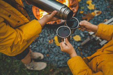 Naklejka premium Close-up people pouring hot drinks from thermos into metal mugs next to campfire. Cozy autumn scene with warm clothing, a woven basket, and the glow of the fire, creating a warm 