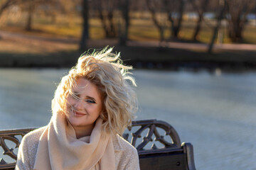 Grown woman sits on bench and smiles at wind ruffling her hair. Windy autumn day in park by water. Copy space