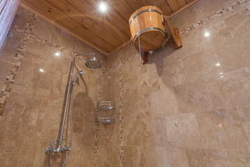 Modern design of the shower in the spa, decorated with beige tiles. A wooden bucket fixed on the wall for pouring cold water. © alhim
