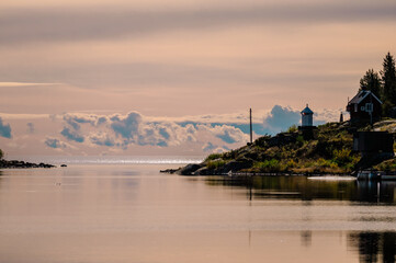 Fototapeta premium A serene scene capturing calm waters reflecting the sky and coastline with a small lighthouse perched on the right. Soft clouds drift in the distance, creating a dreamy atmosphere under soft sunlight