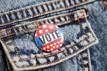 A close-up of a colorful “VOTE” button pinned to a denim jacket, representing political participation and encouragement to vote.