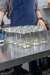 set of empty glass jars lined up on a stainless-steel table in an artisan jam factory. A worker is preparing the jars for filling with handcrafted rosehip jam. 