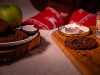 Big flat chocolate chip cookies, candle burning, green apple, warm woolen socks, soft blanket. Person relaxing cozily at home on winter evening. Comfortable lifestyle.