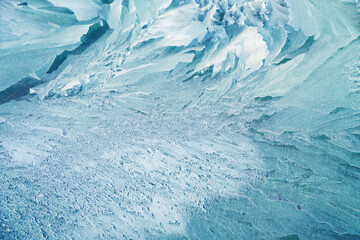 A close-up of ice texture partially covered with snow. The surface displays intricate frozen patterns, with delicate cracks and frosty edges, contrasting the soft, powdery snow scattered across.
