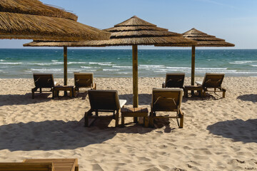 Rows of wooden loungers and straw parasols face the calm, turquoise sea on a sandy beach.