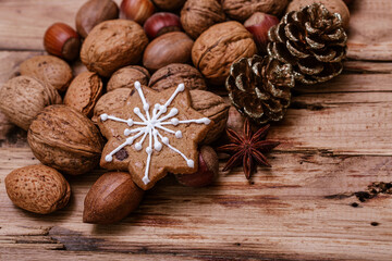 Snowflake-shaped gingerbread cookie with white icing on a wooden table, surrounded by assorted nuts, gold pinecones, and star anise, evoking a warm, festive holiday atmosphere.