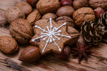 Snowflake-shaped gingerbread cookie with white icing on a wooden table, surrounded by assorted nuts, a gold pinecone, and star anise, creating a cozy, festive holiday atmosphere.