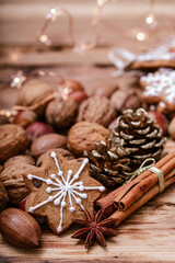 A snowflake-shaped gingerbread cookie with white icing on a wooden table, surrounded by assorted nuts, cinnamon sticks, star anise, a pinecone, and warm star-shaped fairy lights in the background.