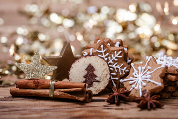 Assorted cookies, including gingerbread stars with white icing and a round cookie with a tree design, on a wooden table with cinnamon sticks, star anise, gold star ornaments, and bokeh lights.
