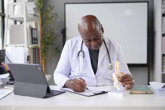 Doctor is smiling while examining an anatomical model of the spine and taking notes on a clipboard in his office - Powered by Adobe