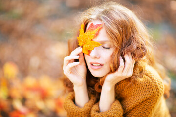 A serene woman with red hair holds an autumn leaf over one eye, eyes closed, wearing a cozy...