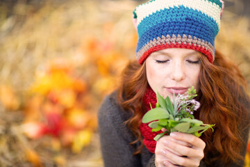 Naklejka premium Serene woman with red hair, wearing a colorful knit hat and gray sweater, holds a small bouquet of flowers close to her face, eyes closed, enjoying the fragrance, surrounded by warm autumn colors.