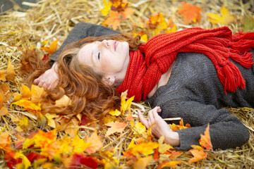 Woman with red hair lies on a bed of straw and colorful autumn leaves, wearing a gray sweater and red scarf, eyes closed, enjoying a peaceful moment surrounded by the warm colors of fall..