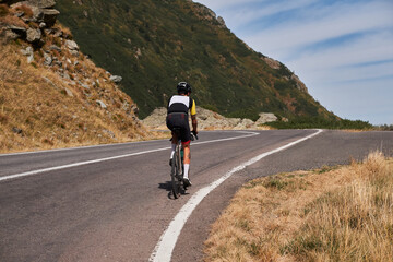 Obraz premium Rear view of a cyclist riding a bike on a winding mountain road. The cyclist is wearing a helmet and cycling gear. The image is a great representation of adventure, freedom, and the beauty of nature.