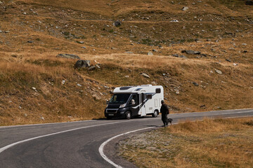 A white camper van driving on a winding road in the mountains, with a person walking nearby. Transfăgărășan road in Romania.

