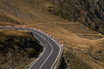 Aerial view of a cyclist riding on a winding asphalt road in the mountains. The road curves through a grassy landscape with rocks and hills, providing a sense of adventure and exploration.