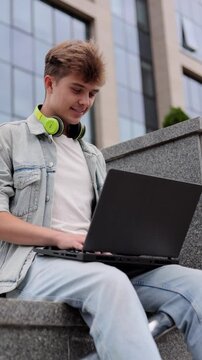Smiling young man student with headphones working on laptop outdoors on a background of university or corporate building. Vertical video.