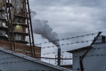 A large industrial facility releases smoke into the gray sky, showcasing the complexity of urban life amid towering structures and barbed wire