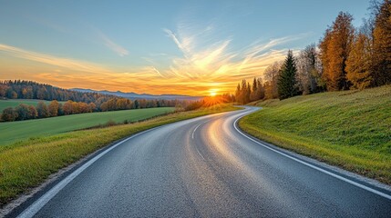 Obraz premium A winding asphalt road leads through a scenic countryside landscape, with golden sunset rays illuminating the horizon.