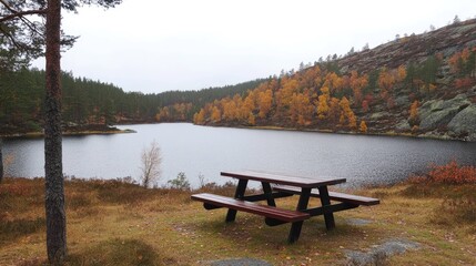 Obraz premium A wooden picnic table sits overlooking a lake with fall foliage on the hillside.