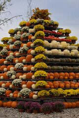 Pyramid-shaped display of pumpkins and colorful flowers arranged in pots for an autumn harvest festival on a cloudy day. Concept of seasonal decoration, fall harvest and festive outdoor events