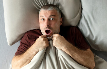 Stressed scared European middle aged man lying in bed and holding blanket, looking at camera with...