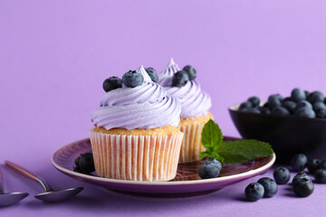Plate of tasty cupcakes with blueberries on lilac background