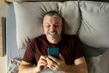 Technology and people concept. Happy smiling middle aged man texting on smartphone lying in bed at home in the morning, above view