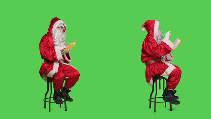 Father christmas reading literature in studio with full body greenscreen, enjoying hobby with fiction or poetry book. Wise cultured saint nick reads novel story for lecture, having fun.