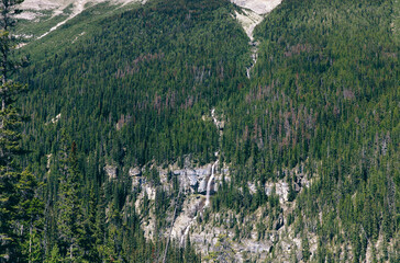 A lush green forest covers a mountain slope with a series of small waterfalls cascading down,...