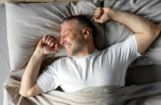 Top view of happy middle aged man enjoying happy morning, lying in bed and stretching his body after waking up, feeling well-rested