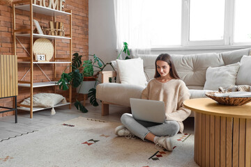 Young woman using laptop on carpet near beige sofa in living room