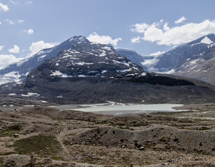 A rocky and rugged terrain with a clear lake in the center and a breathtaking snow-capped mountain range in the background under a bright, clear sky.