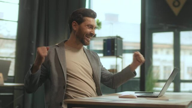 Happy young Caucasian man sitting at desk with laptop celebrating success. Excited overjoyed stylish businessman at workplace in office. Tracking moving around. 360 degree shot. Speed. Cinematic