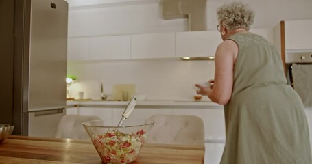 An elderly woman is preparing a salad in her modern kitchen. She is adding ingredients to a bowl.