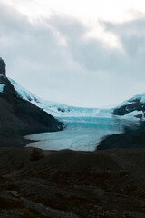 A glacier flows through a mountain valley under an overcast, cloudy sky, capturing the tranquil yet powerful presence of ice as it carves through the rugged landscape.